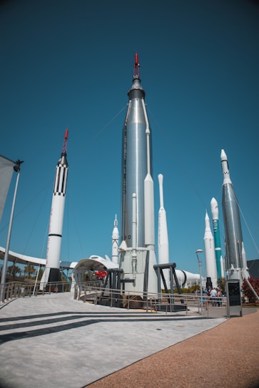 A collection of large, metallic rockets standing upright against a clear blue sky, with some red and black detailing. The area appears to be an outdoor exhibition, with visitors walking around and observing the display. There is a concrete path and platforms surrounding the rockets, and some greenery can be seen in the background.
