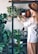 A person with long blonde hair and a brown hat tends to various houseplants on a shelving unit. The shelves are filled with a diverse collection of potted plants, featuring lush green leaves. Natural light softly illuminates the scene, creating a tranquil, earthy ambiance.