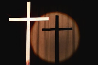 A wooden cross is illuminated in the foreground against a dark background. The shadow of the cross is cast onto a circular spot of light on a curtain.