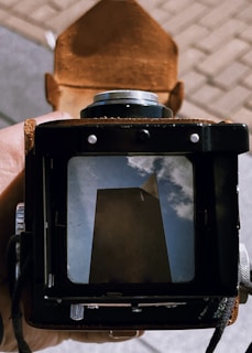 A vintage camera viewfinder captures an artistic reflection of a tall, dark building against a backdrop of blue sky and clouds. The image is framed by the camera's matte black and brown leather components, creating a nostalgic and artistic feel.