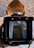 A vintage camera viewfinder captures an artistic reflection of a tall, dark building against a backdrop of blue sky and clouds. The image is framed by the camera's matte black and brown leather components, creating a nostalgic and artistic feel.