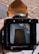 A vintage camera viewfinder captures an artistic reflection of a tall, dark building against a backdrop of blue sky and clouds. The image is framed by the camera's matte black and brown leather components, creating a nostalgic and artistic feel.