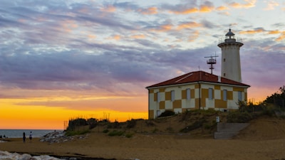 A lighthouse stands beside a sandy beach as the sun sets on the horizon, casting vibrant hues of orange and purple across the sky. The building is characterized by a red roof and a checkered yellow and beige facade. Two people are visible walking near the shoreline.