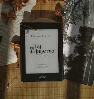 A Kindle displaying the title 'O Rei do Inverno' by Bernard Cornwell is placed on a bamboo mat. Surrounding the Kindle are dried leaves, some loose book pages, and black-and-white photographs, creating a vintage and cozy atmosphere.
