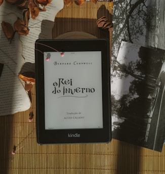 A Kindle displaying the title 'O Rei do Inverno' by Bernard Cornwell is placed on a bamboo mat. Surrounding the Kindle are dried leaves, some loose book pages, and black-and-white photographs, creating a vintage and cozy atmosphere.