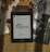 A Kindle displaying the title 'O Rei do Inverno' by Bernard Cornwell is placed on a bamboo mat. Surrounding the Kindle are dried leaves, some loose book pages, and black-and-white photographs, creating a vintage and cozy atmosphere.