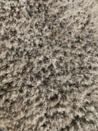 A close-up view of a textured surface with dense, tightly packed curls resembling wool or fur. The color palette is composed of varying shades of brown and beige, creating a natural and warm appearance.