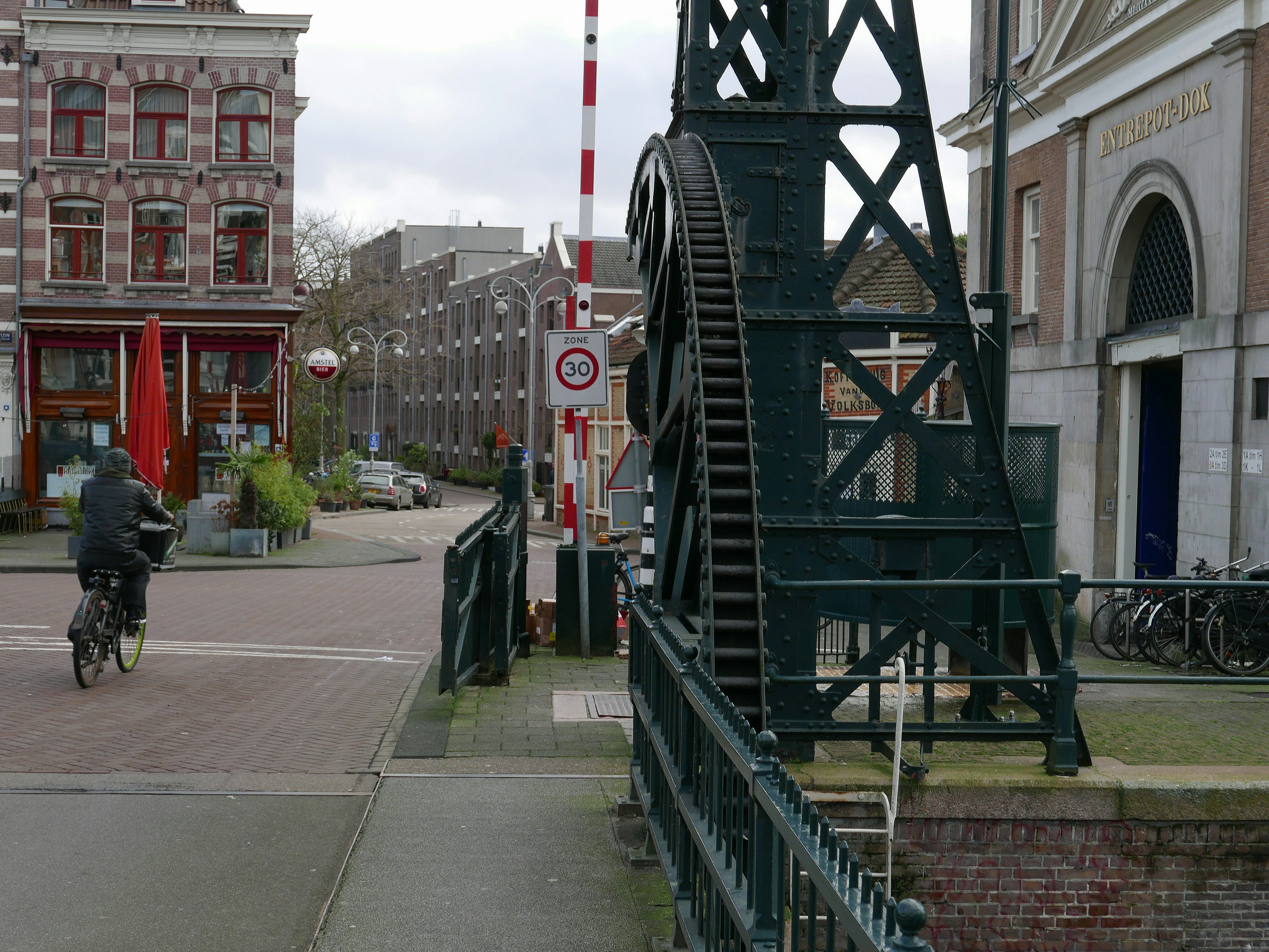 A man is biking over the drawbridge in the old city van Amsterdam. It is a historical place here with to the right the entrance gate of the large Entrepotdok and the long row store-house buildings. All grey texture of bricks and old architecture of c. 1810 in this street view. Free pic of Amsterdam streets and roads - Dutch street photography in free photo by Fons Heijnsbroek, Netherlands.