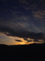 The image depicts a dramatic landscape with dark hills in the foreground silhouetted against a vibrant sunset. The sky is filled with dark clouds, with streaks of warm sunlight breaking through, casting a golden hue on the horizon. The scene conveys a sense of calm and beauty as day transitions into night.
