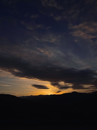 The image depicts a dramatic landscape with dark hills in the foreground silhouetted against a vibrant sunset. The sky is filled with dark clouds, with streaks of warm sunlight breaking through, casting a golden hue on the horizon. The scene conveys a sense of calm and beauty as day transitions into night.