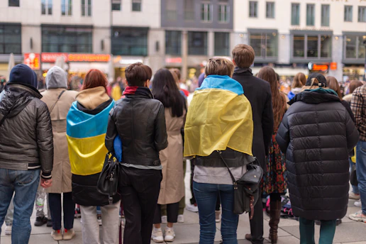 A group of people standing with their backs to the camera, with some wearing flags. They are gathered in an urban setting with buildings and a street in the background. The atmosphere seems to be social or possibly political.