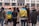 A group of people standing with their backs to the camera, with some wearing flags. They are gathered in an urban setting with buildings and a street in the background. The atmosphere seems to be social or possibly political.