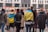 A group of people standing with their backs to the camera, with some wearing flags. They are gathered in an urban setting with buildings and a street in the background. The atmosphere seems to be social or possibly political.