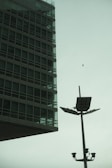 A modern glass and steel building with grid-like windows is positioned on the left side, juxtaposed against a tall lamppost with solar panels and surveillance cameras. A solitary bird is captured mid-flight against the overcast sky.