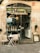 An inviting storefront of a hair boutique, featuring a glass door and window displaying hair products neatly arranged inside. The exterior includes two cushioned chairs and a small round table with a tea set, creating a welcoming atmosphere. The building's facade is a combination of stone and decorative metalwork.