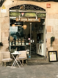 An inviting storefront of a hair boutique, featuring a glass door and window displaying hair products neatly arranged inside. The exterior includes two cushioned chairs and a small round table with a tea set, creating a welcoming atmosphere. The building's facade is a combination of stone and decorative metalwork.