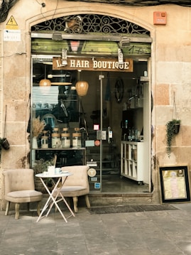 An inviting storefront of a hair boutique, featuring a glass door and window displaying hair products neatly arranged inside. The exterior includes two cushioned chairs and a small round table with a tea set, creating a welcoming atmosphere. The building's facade is a combination of stone and decorative metalwork.