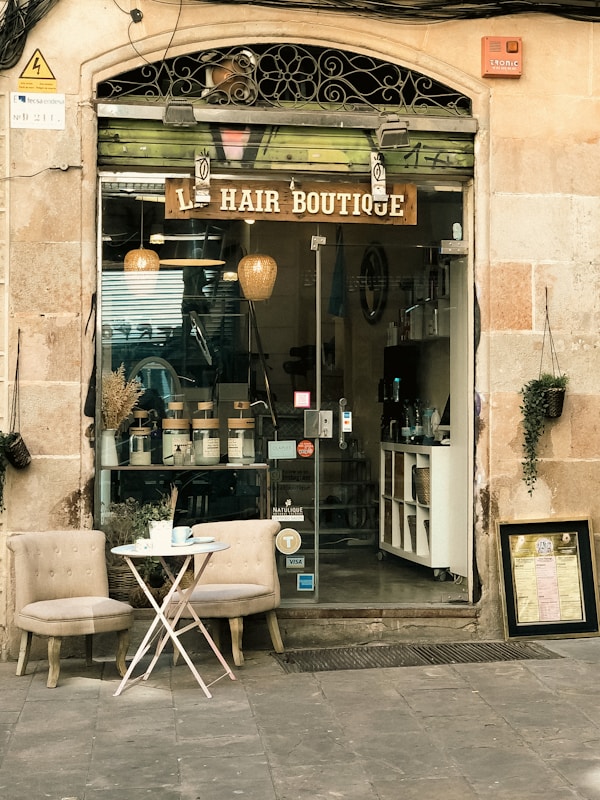 An inviting storefront of a hair boutique, featuring a glass door and window displaying hair products neatly arranged inside. The exterior includes two cushioned chairs and a small round table with a tea set, creating a welcoming atmosphere. The building's facade is a combination of stone and decorative metalwork.