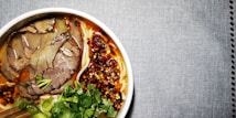 A bowl filled with sliced beef, noodles, cilantro, green onions, and a spicy red chili oil sauce. The broth appears rich and flavorful, with the vibrant colors of the herbs and chili oil creating an appetizing presentation. The bowl is set against a textured gray background.