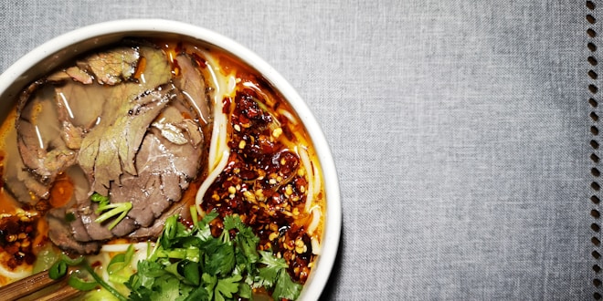 A bowl filled with sliced beef, noodles, cilantro, green onions, and a spicy red chili oil sauce. The broth appears rich and flavorful, with the vibrant colors of the herbs and chili oil creating an appetizing presentation. The bowl is set against a textured gray background.