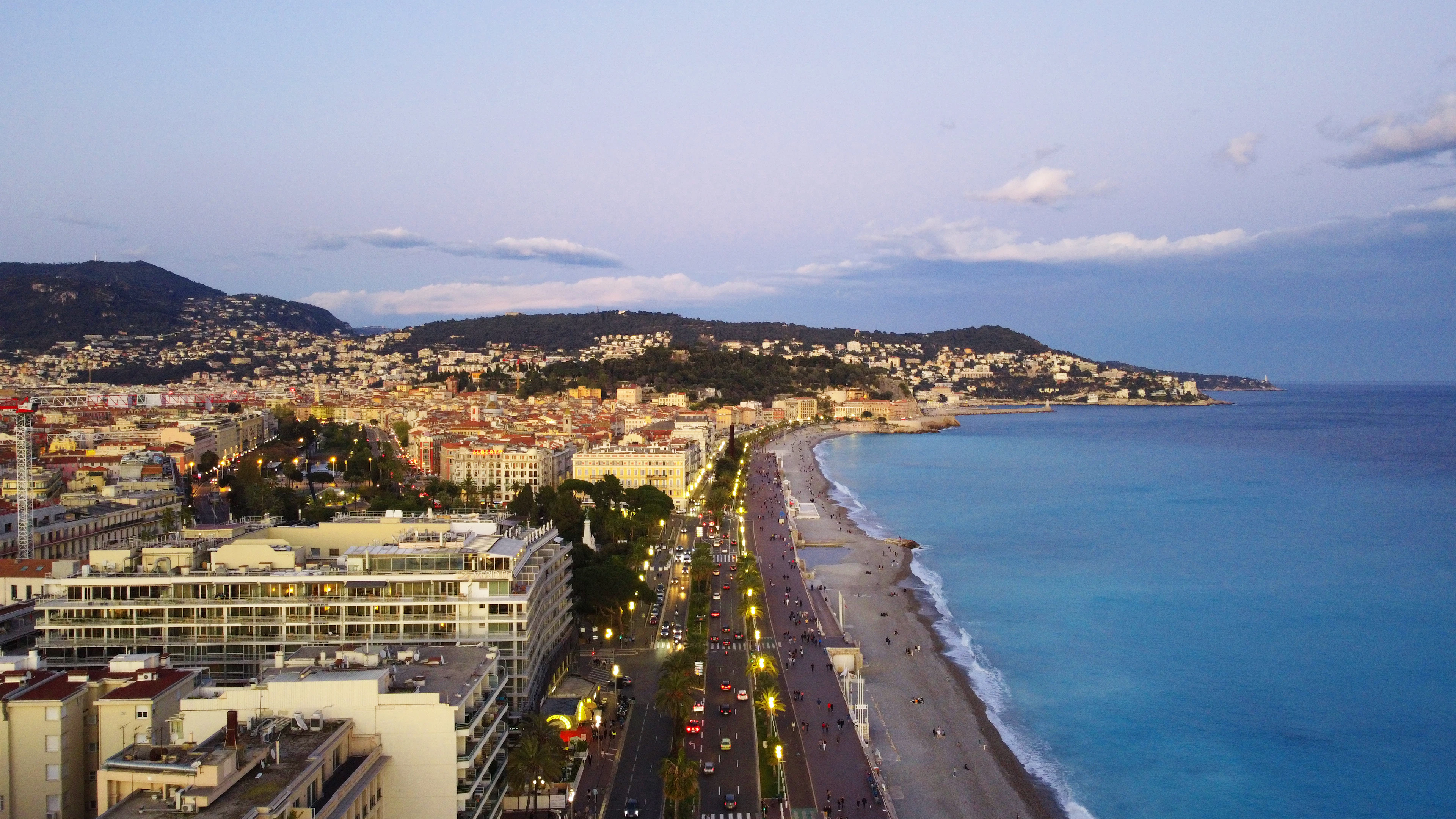 Aerial view of the curved Promenade des Anglais and Baie des Anges at dusk with city lights coming on.
