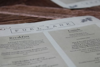 A restaurant menu titled 'The Fuel Tank' features sections for Breakfast and Lunch. It offers a Full English Breakfast and various lunch options, including sandwiches. The menu includes details on serving times and pricing. The menu page is laid over a wooden surface.