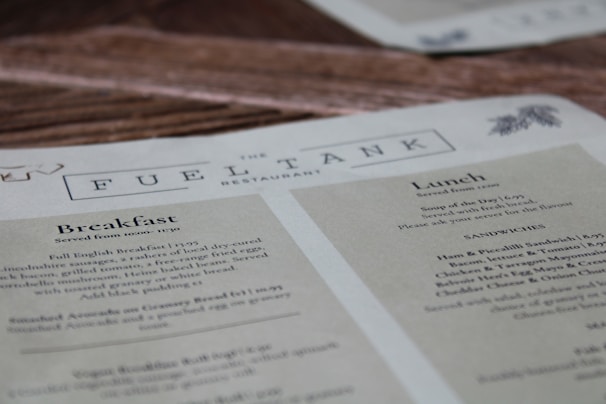 A restaurant menu titled 'The Fuel Tank' features sections for Breakfast and Lunch. It offers a Full English Breakfast and various lunch options, including sandwiches. The menu includes details on serving times and pricing. The menu page is laid over a wooden surface.