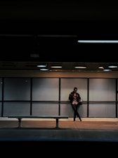 A person stands alone in a dimly lit subway station, leaning against a perforated metal wall while looking at a smartphone. The empty bench in the foreground emphasizes solitude. Overhead lights cast a subtle glow, creating a moody atmosphere.