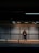 A person stands alone in a dimly lit subway station, leaning against a perforated metal wall while looking at a smartphone. The empty bench in the foreground emphasizes solitude. Overhead lights cast a subtle glow, creating a moody atmosphere.