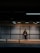 A person stands alone in a dimly lit subway station, leaning against a perforated metal wall while looking at a smartphone. The empty bench in the foreground emphasizes solitude. Overhead lights cast a subtle glow, creating a moody atmosphere.