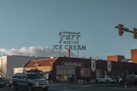 A vintage ice cream shop is situated on a street corner, featuring a large retro-style sign that reads 'Farr Better Ice Cream.' The building is constructed with red bricks and has a classic, nostalgic appearance. Several cars are parked nearby, and a traffic light is visible above the intersection.