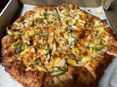 A freshly baked pizza topped with slices of pineapple, green bell peppers, cheese, and bits of cooked chicken. The crust is golden brown and slightly thick. The pizza is placed on a white paper inside a cardboard delivery box.