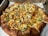 A freshly baked pizza topped with slices of pineapple, green bell peppers, cheese, and bits of cooked chicken. The crust is golden brown and slightly thick. The pizza is placed on a white paper inside a cardboard delivery box.