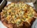 A freshly baked pizza topped with slices of pineapple, green bell peppers, cheese, and bits of cooked chicken. The crust is golden brown and slightly thick. The pizza is placed on a white paper inside a cardboard delivery box.