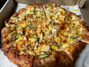 A freshly baked pizza topped with slices of pineapple, green bell peppers, cheese, and bits of cooked chicken. The crust is golden brown and slightly thick. The pizza is placed on a white paper inside a cardboard delivery box.
