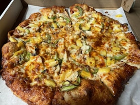 A freshly baked pizza topped with slices of pineapple, green bell peppers, cheese, and bits of cooked chicken. The crust is golden brown and slightly thick. The pizza is placed on a white paper inside a cardboard delivery box.