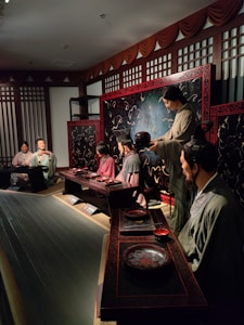 A group of figures dressed in traditional East Asian attire are seated around a low table, engaging in what appears to be a historical reenactment or display. The setting features ornate wooden screens with intricate patterns and a dimly lit, warm atmosphere.