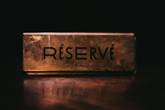 A gold-colored reserved sign sits prominently on a table surface, with the word 'RÉSERVÉ' in bold, black lettering. The background is dark, adding emphasis to the sign.