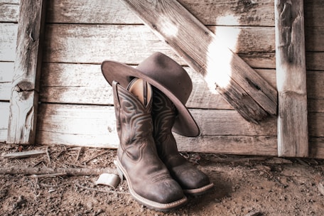 Close-up of vintage leather boots and a worn cowboy hat resting on old wooden fence rails.