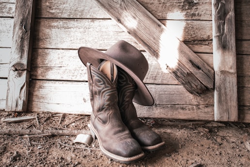 A pair of cowboy boots and a cowboy hat are placed against a rustic wooden wall. The wooden planks have a weathered appearance, creating a vintage and Western vibe. The boots and hat are positioned on dirt-covered ground, adding to the rustic atmosphere.
