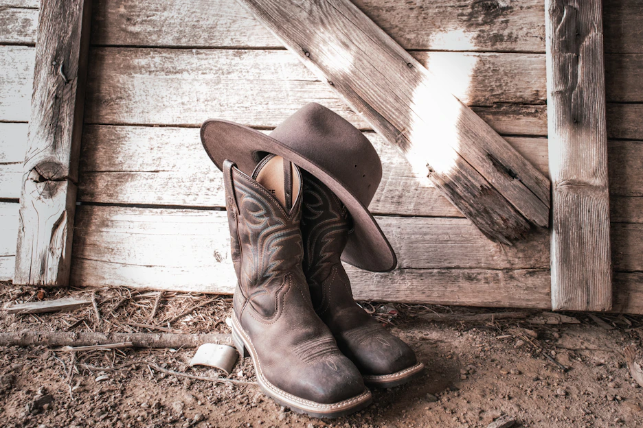 Rustic cowboy boots and leather jacket displayed on a wooden fence with a sunset background