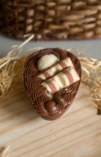 A chocolate dessert shaped like an egg with intricate swirls of chocolate on top. It features cream-filled wafer sticks and a small white chocolate piece as toppings. The dessert is presented on a light wooden surface with straw and a wicker basket in the background.