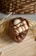 A chocolate dessert shaped like an egg with intricate swirls of chocolate on top. It features cream-filled wafer sticks and a small white chocolate piece as toppings. The dessert is presented on a light wooden surface with straw and a wicker basket in the background.