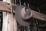 A cowboy hat hangs on a wooden beam inside a rustic wooden structure, with visible rough textures and natural tones.