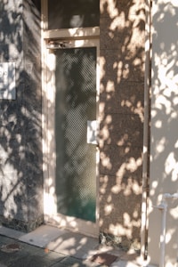 A glass door with a textured pattern is set within a stone or marble-walled exterior. Shadows of tree leaves are cast on the wall and door, creating a natural and textured interplay of light. The scene is tranquil, featuring a combination of man-made and natural elements.