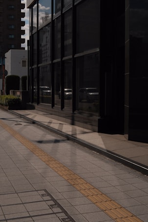 Reflective glass windows line a modern building on a city street, capturing shadows of nearby structures. The pavement is marked with a textured, yellow strip to aid visually impaired pedestrians. Surrounding buildings, some residential, add to the urban atmosphere, with soft shadows cast by the afternoon sun.