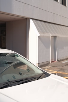 A partial view of a white car parked near a beige building with a minimalist facade. The building has a small door and windows, with a clean, modern architectural style. The scene is brightly lit by sunlight, creating clear shadows on the surfaces.