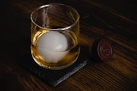 A glass of whiskey with a large spherical ice cube rests on a dark stone coaster, placed on a wooden surface. Beside it is a round wooden object with an engraved design.