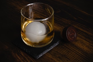 A glass of whiskey with a large spherical ice cube rests on a dark stone coaster, placed on a wooden surface. Beside it is a round wooden object with an engraved design.
