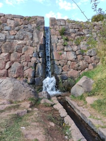 A narrow waterfall cascades down a rustic stone wall, surrounded by greenery. The stones are arranged in a rough yet organized manner, providing a textured backdrop. Water flows into a small channel at the bottom, continuing the serene environment.